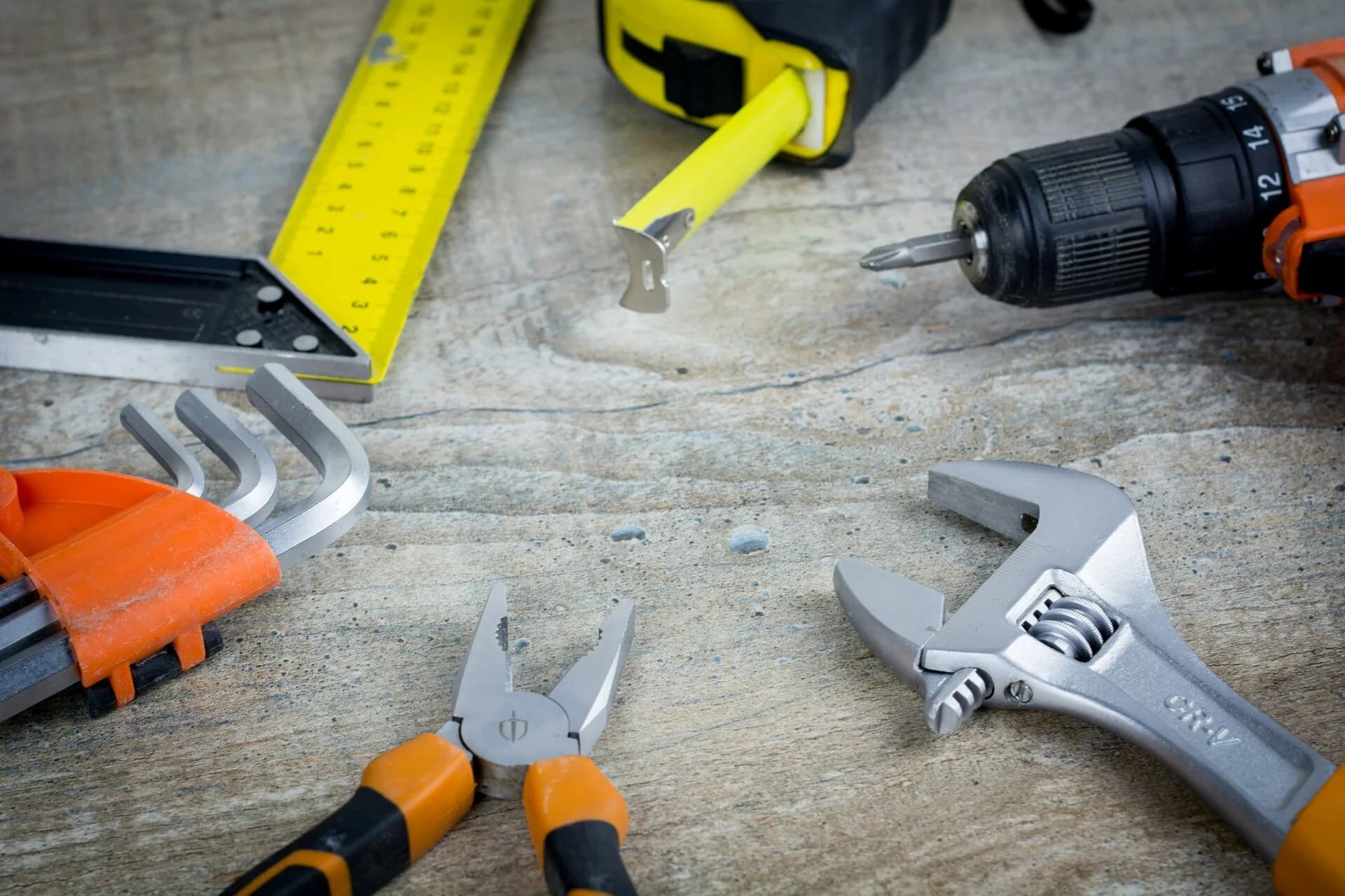 A tape measure, drill, pliers and wrench on a worn workbench
